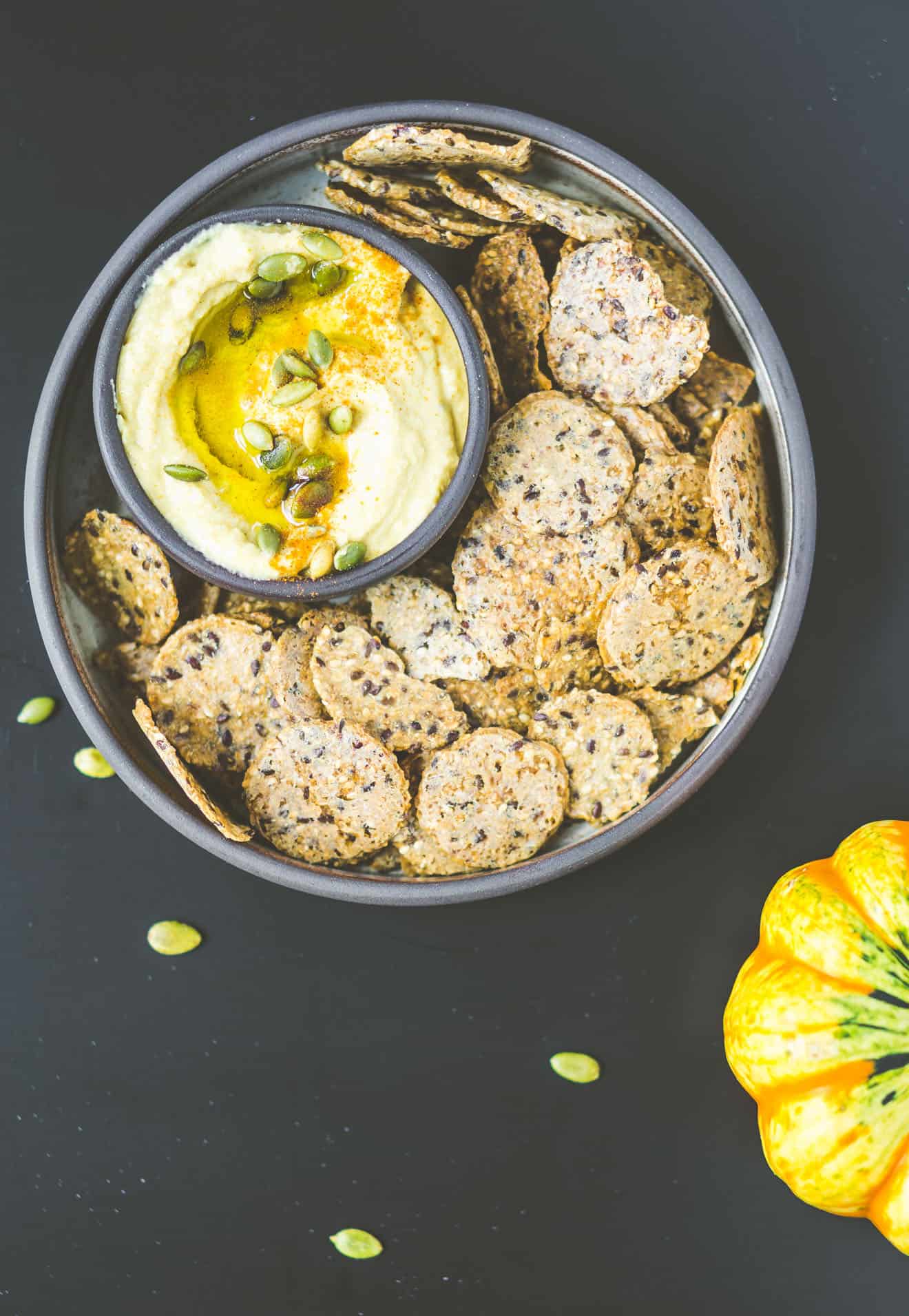 A face down view of hummus with pumpkin seeds inside a bowl with crackers. 