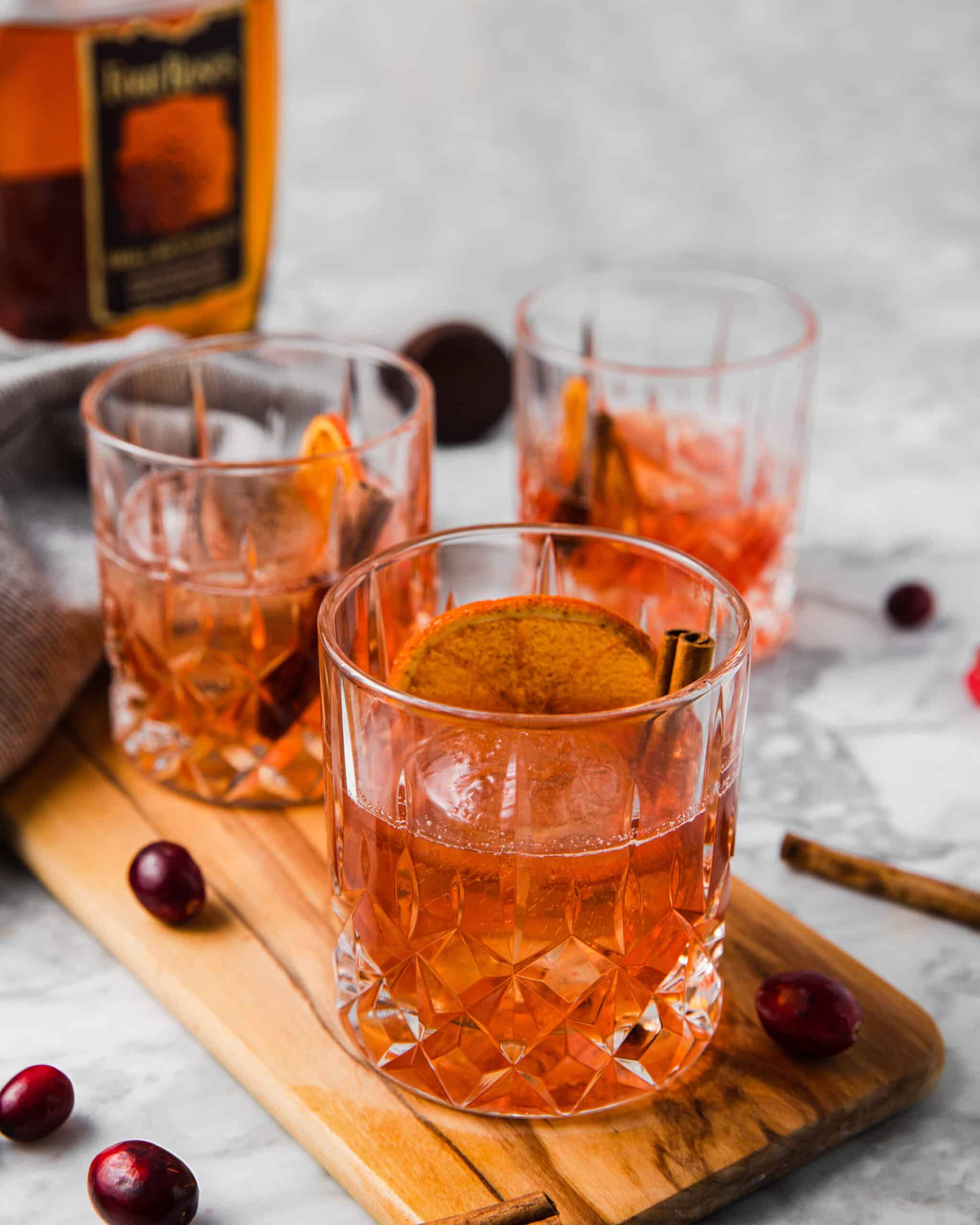 3 glasses of cranberry old fashioned with a orange wheel and cinnamon stick.