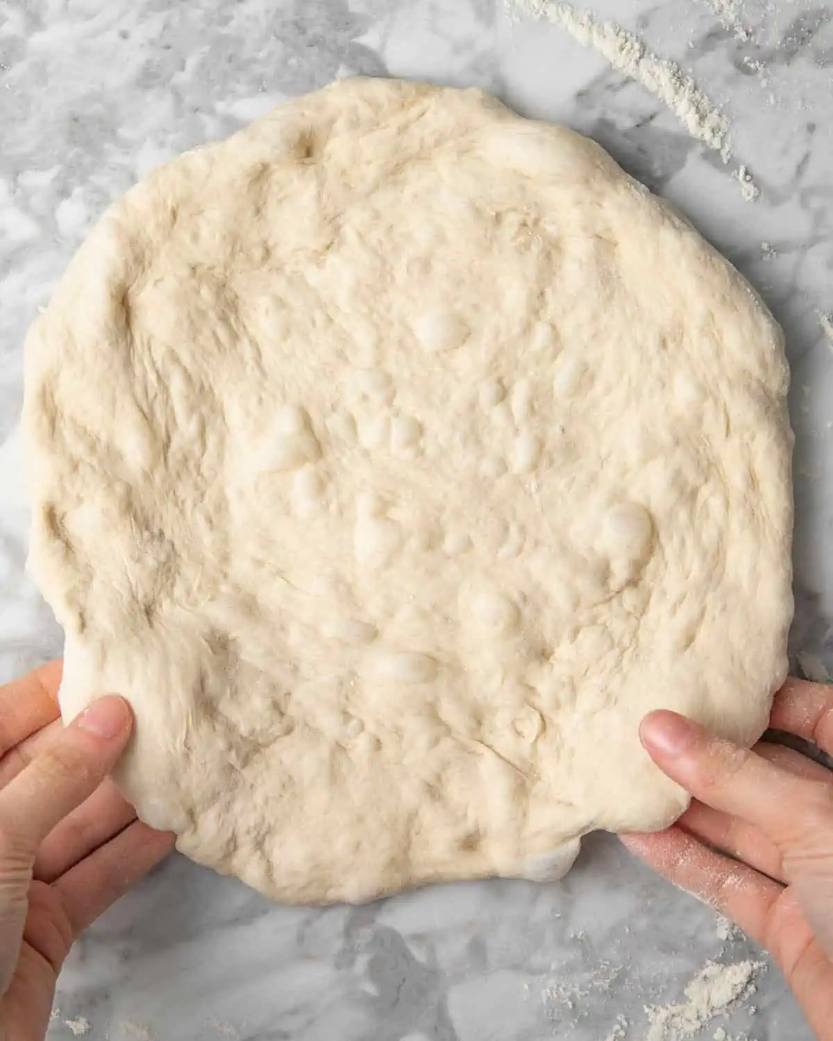 two hands shaping pizza dough on a white marble counter.
